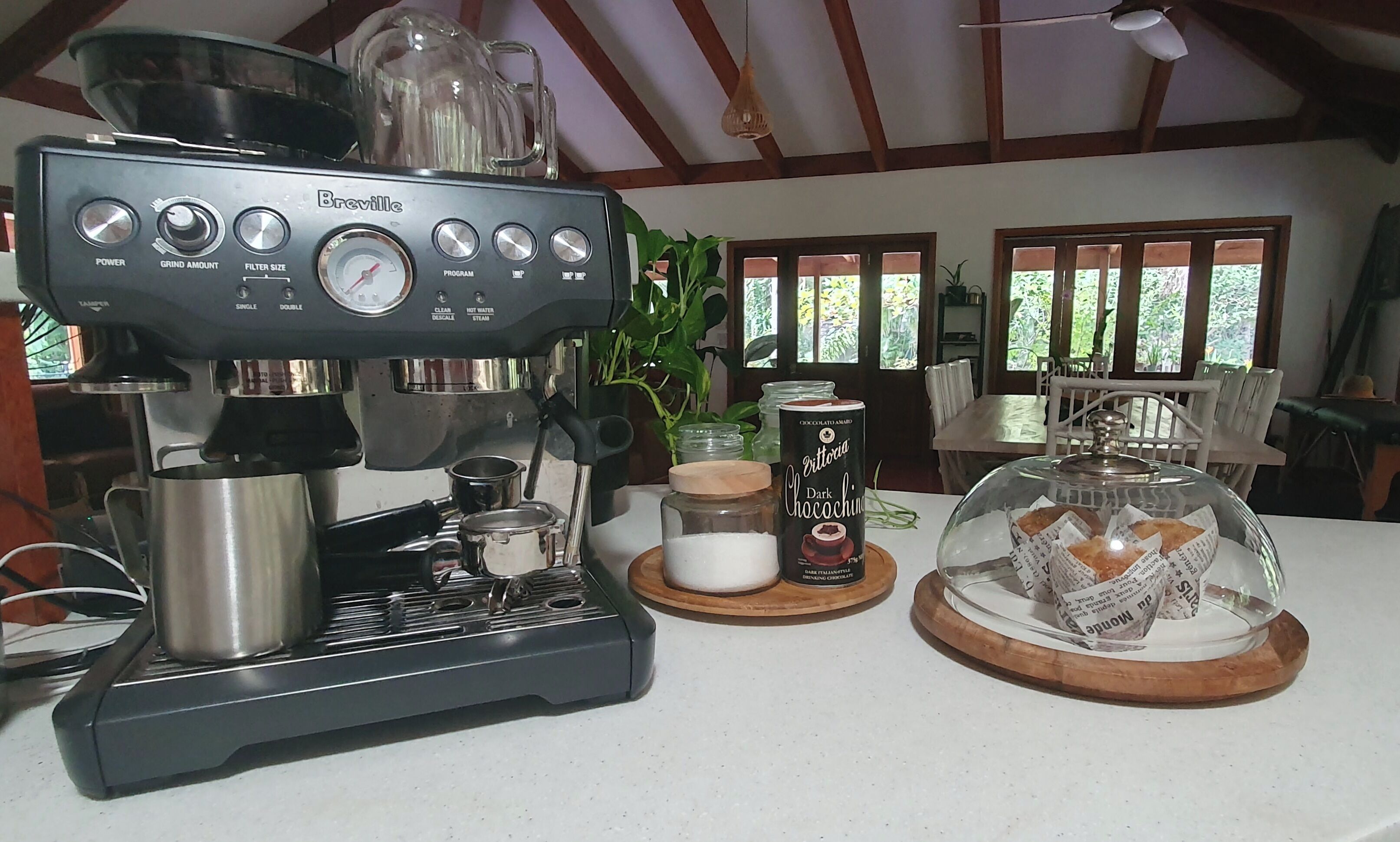 A coffee machine with a grinder, a jar of chocolate syrup, and a glass dome with pastries on a countertop.