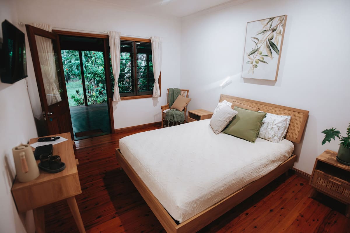 A bedroom with a wooden bed, a small bedside table, a chair, a window with curtains, and a door leading to an outdoor area.