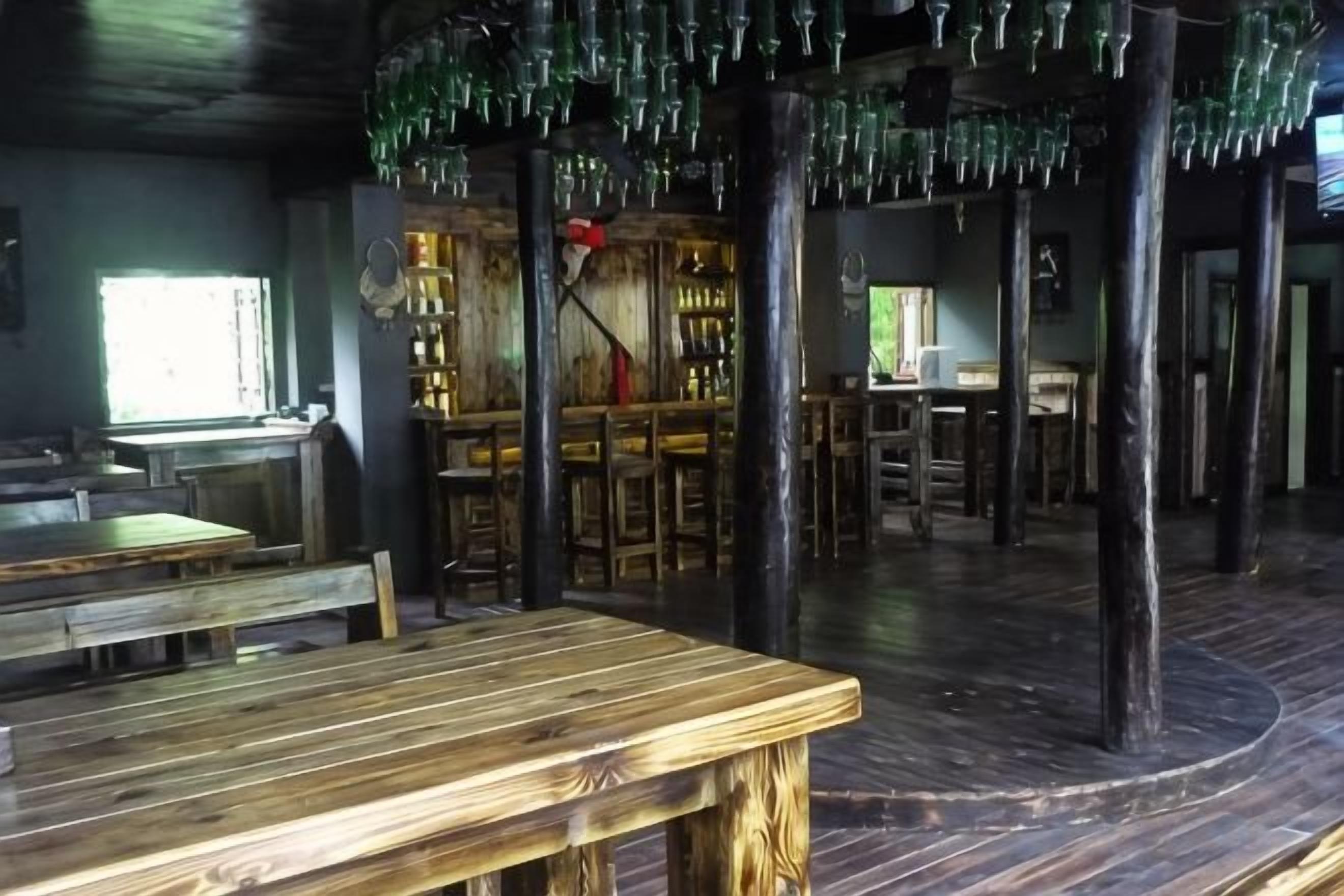 Un intérieur rustique avec des tables et des chaises en bois, un coin bar et un plafond original orné de bouteilles en verre suspendues.