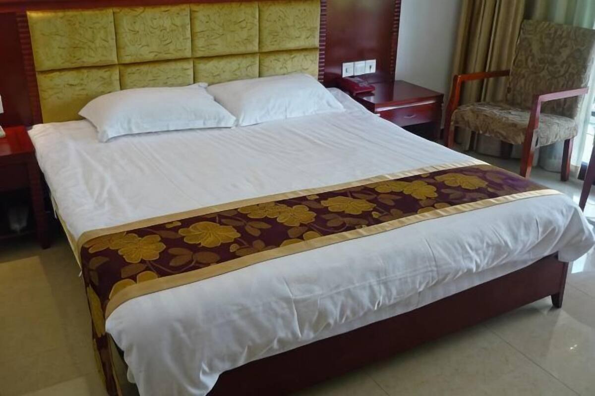 Una cama bien hecha con una colcha estampada con motivos florales, un cabecero de madera, una mesita de noche con una lámpara y una silla.