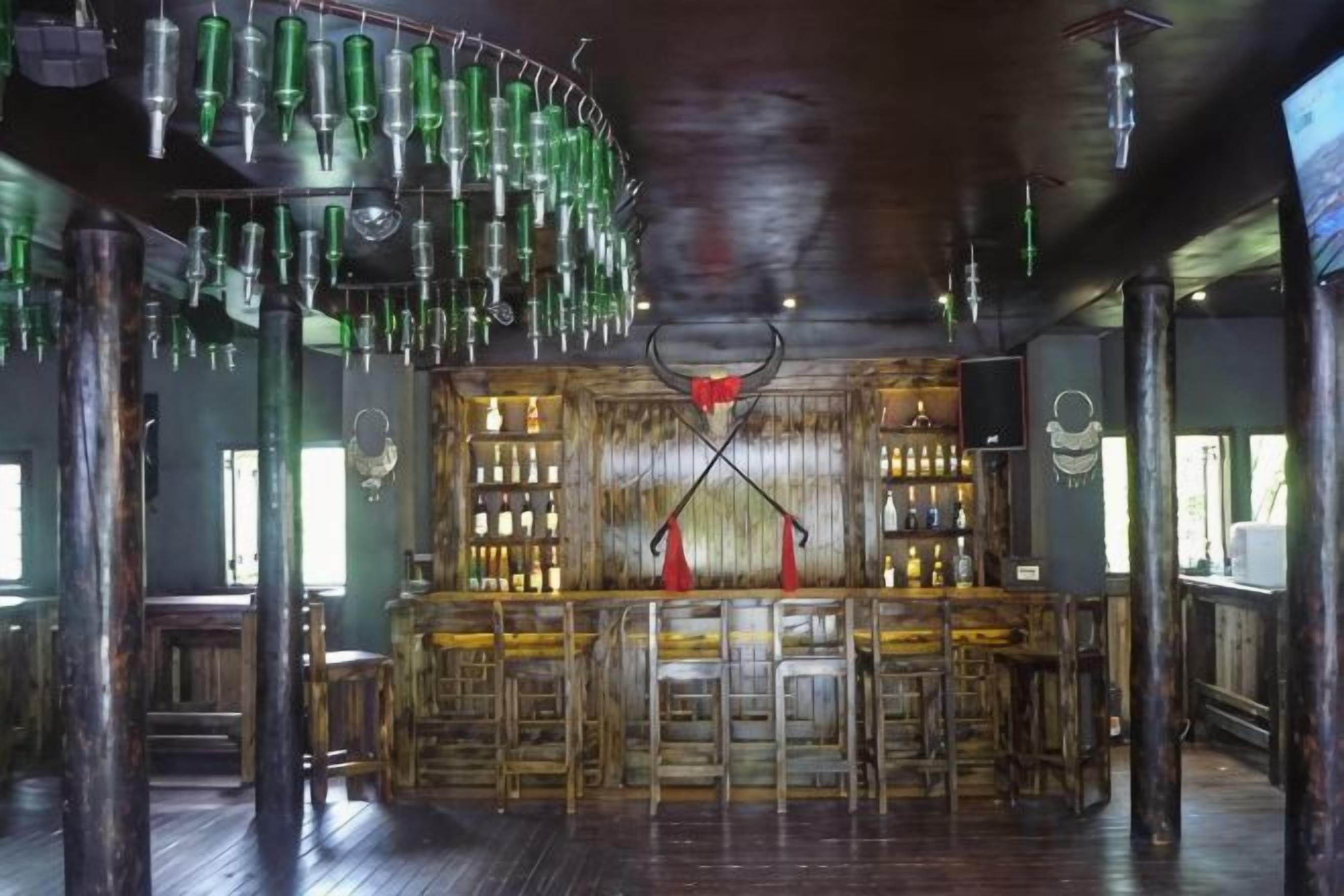 A bar area with a wooden bar counter, stools, and hanging green bottles.