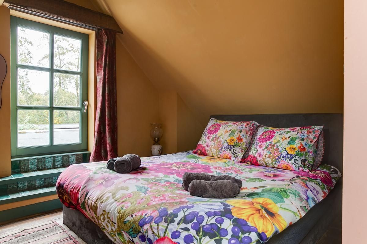 Een slaapkamer met een groot bed, bloemenbeddengoed, een raam met gordijnen en uitzicht op het omliggende landschap.
