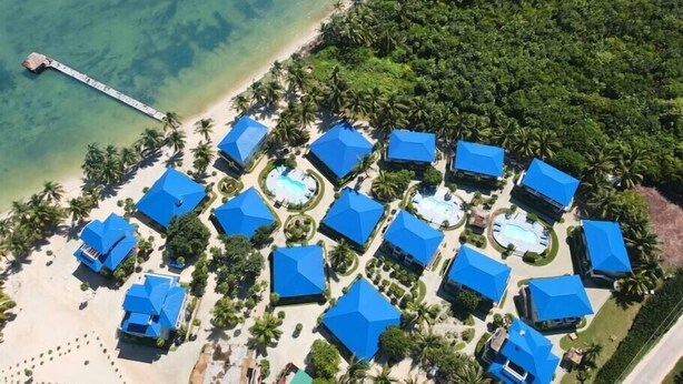 An aerial view of a resort with multiple blue-roofed buildings, a beach, and a pier extending into the water. - 20 Steps From The Beach @ Sapphire Beach Resort (San Pedro Town)