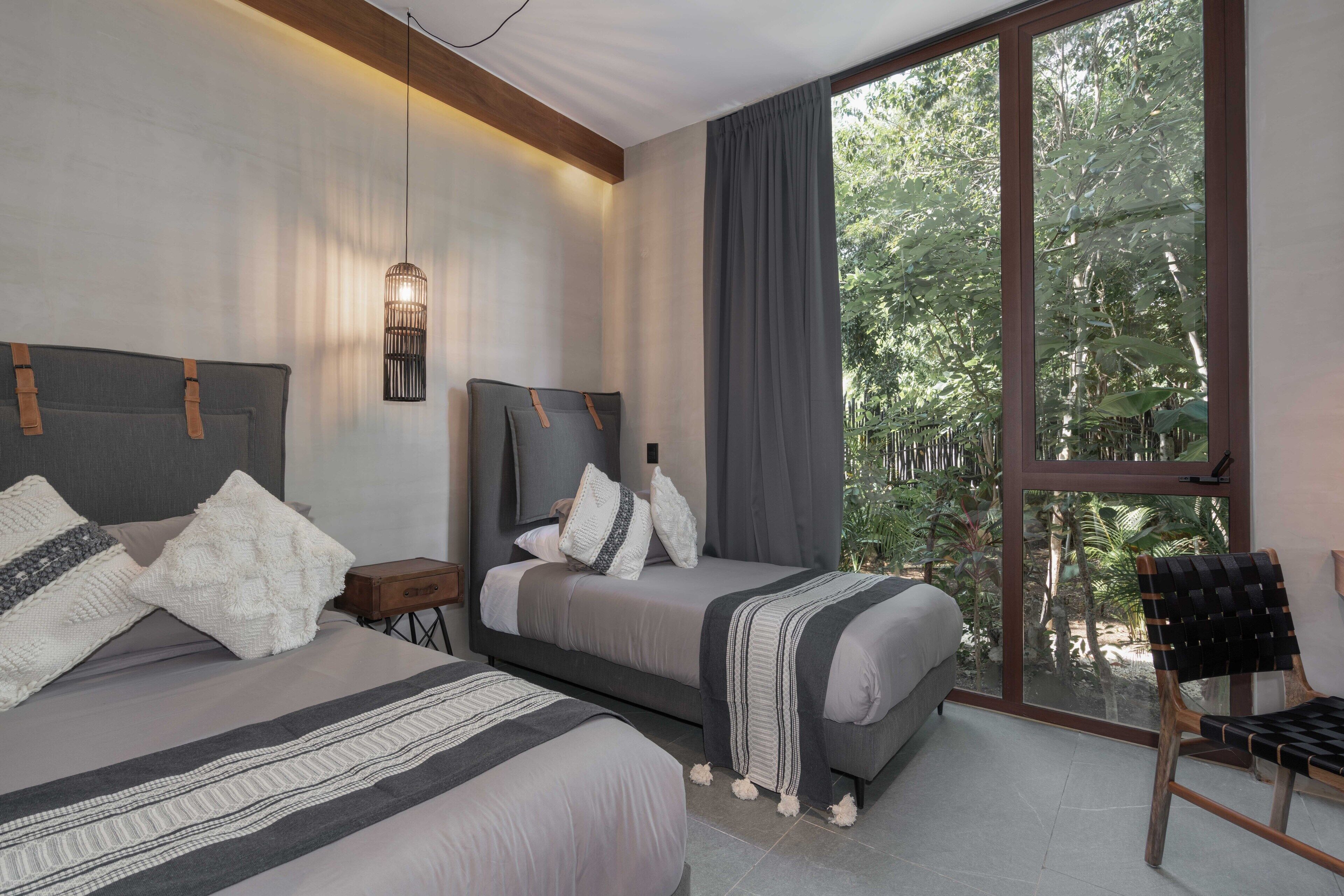 A bedroom with two beds, a large window with a view of greenery, a hanging light fixture, and a chair.