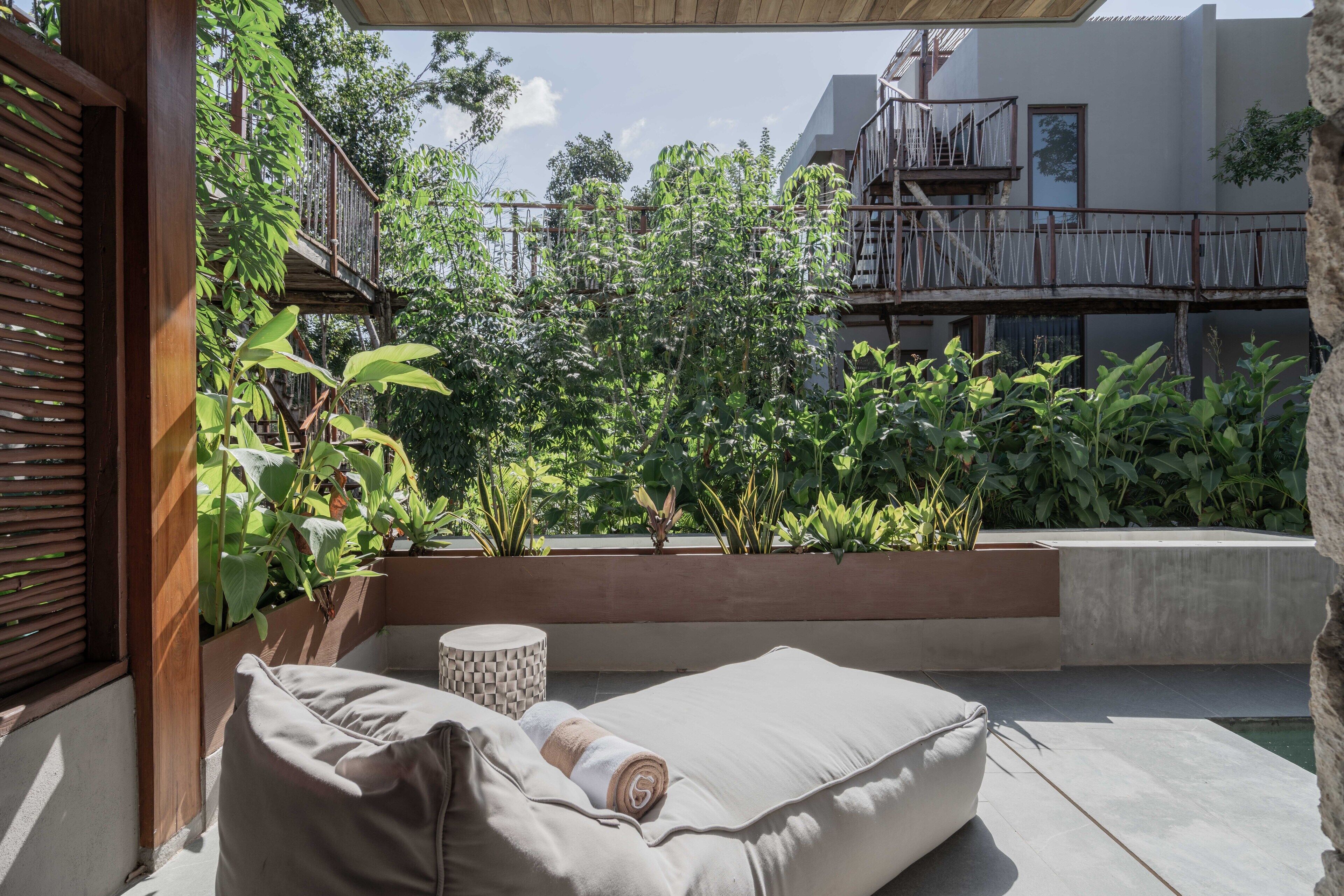 Un espacio al aire libre moderno con un banco acolchado y una mesa redonda, rodeado de abundante follaje y una terraza de madera.