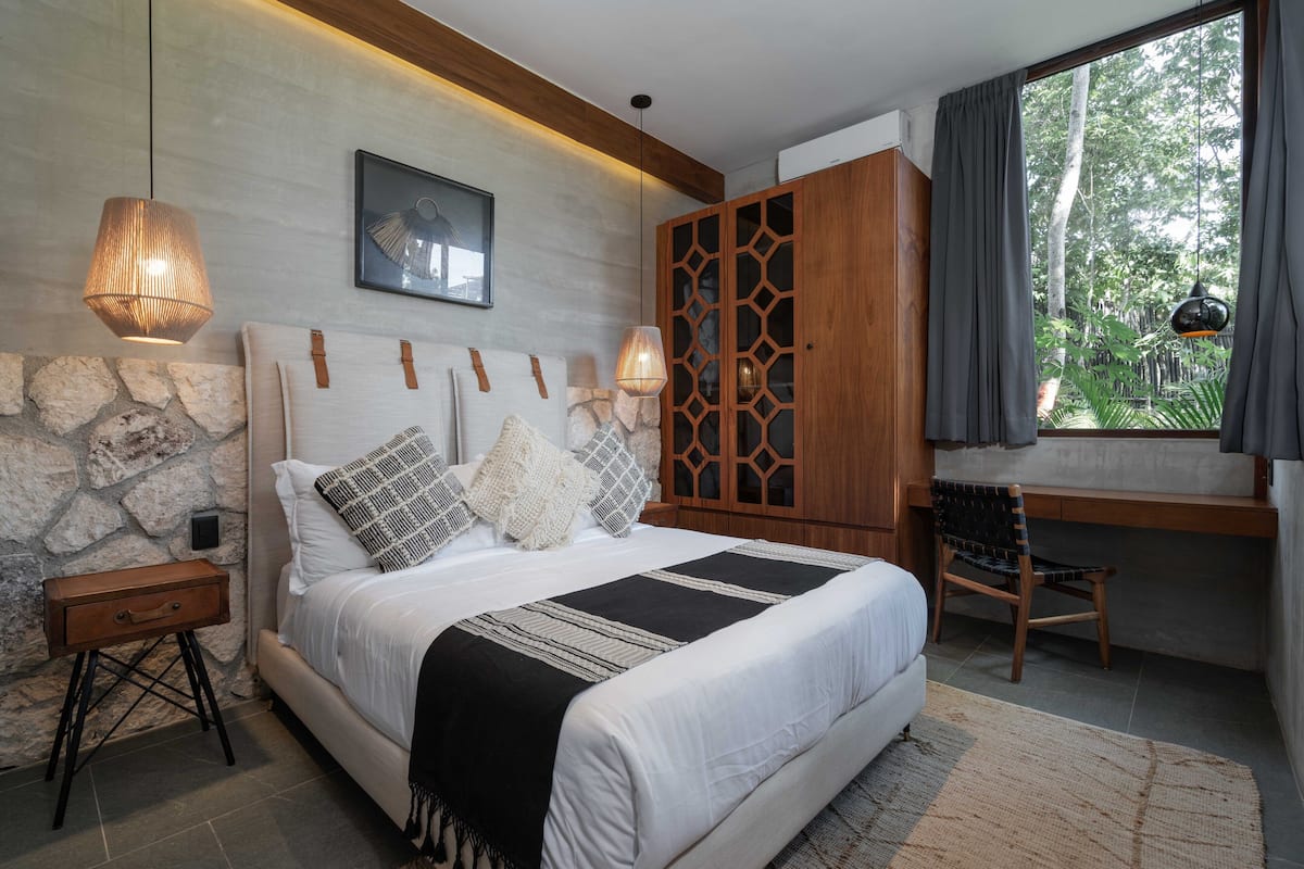 A bedroom with a large bed, a wooden wardrobe, a desk, and a window with a view of greenery.