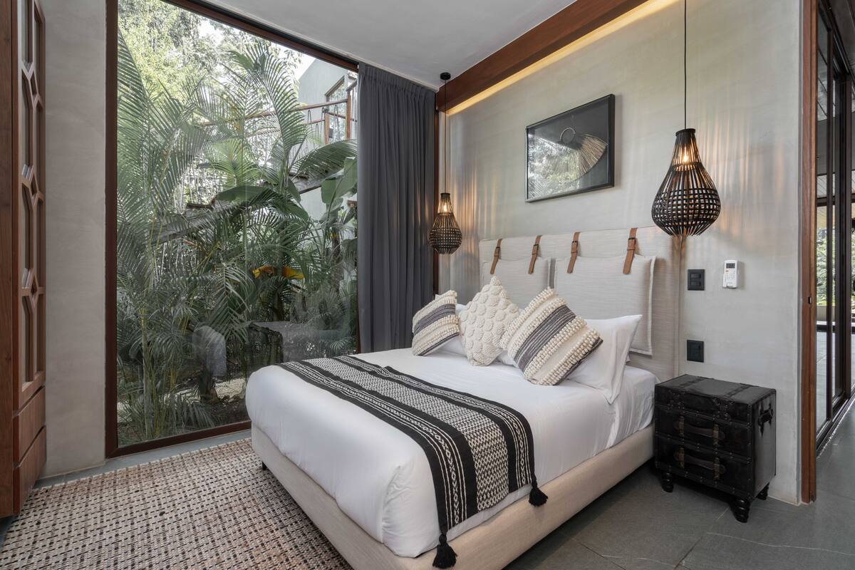 A bedroom with a large sliding glass door leading to a tropical garden, a bed with a white headboard, and two wall-mounted lamps.