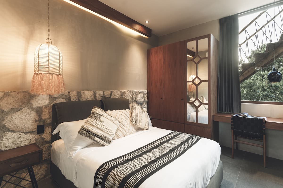 A bedroom with a stone wall, a bed with white bedding, a hanging light fixture, and a view of a wooden staircase.