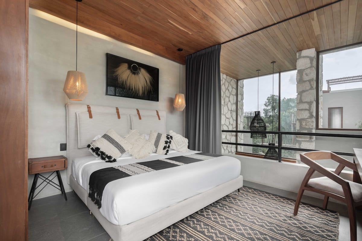 A modern bedroom with a large bed, a wooden headboard, a mounted artwork, and a view of the outdoors through a balcony.