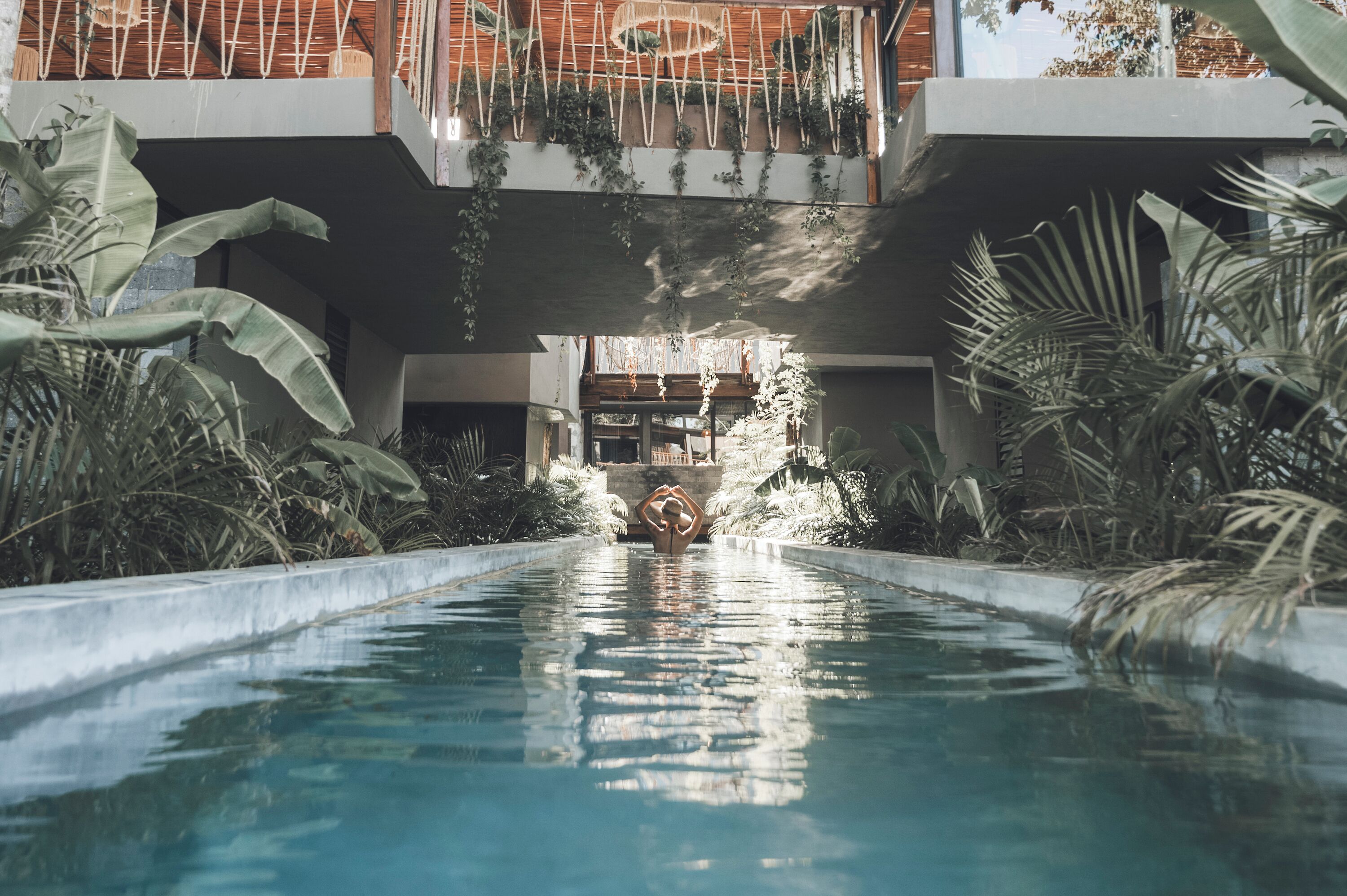 A swimming pool with a central statue, surrounded by lush greenery and a modern building structure with balconies.