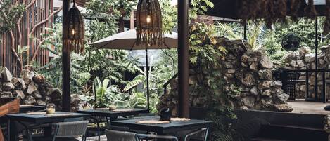 An outdoor dining area with tables and chairs, surrounded by greenery and stone walls.