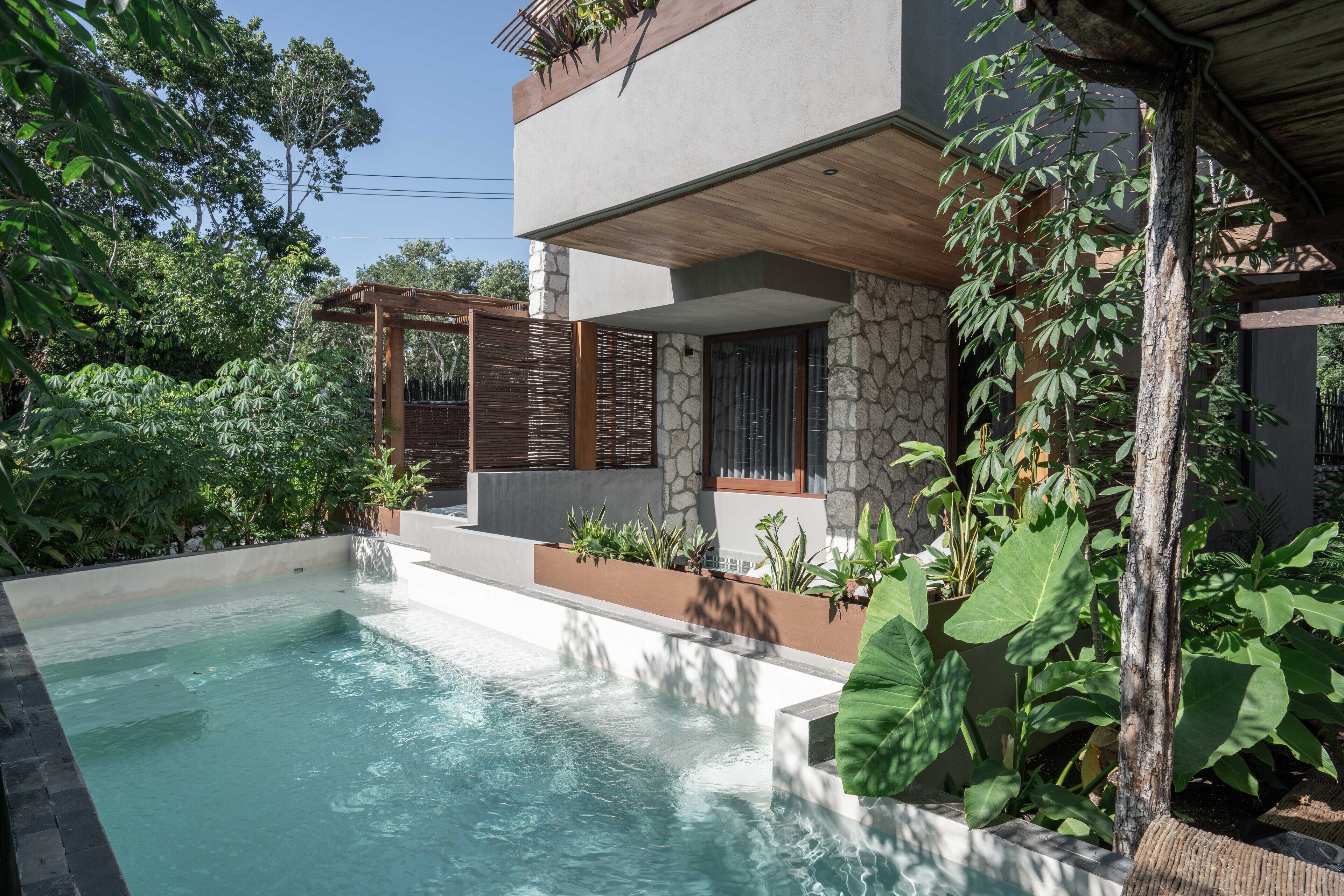 Uma área de piscina moderna ao ar livre, com design em pedra e madeira, rodeada por vegetação exuberante e um pergolado.