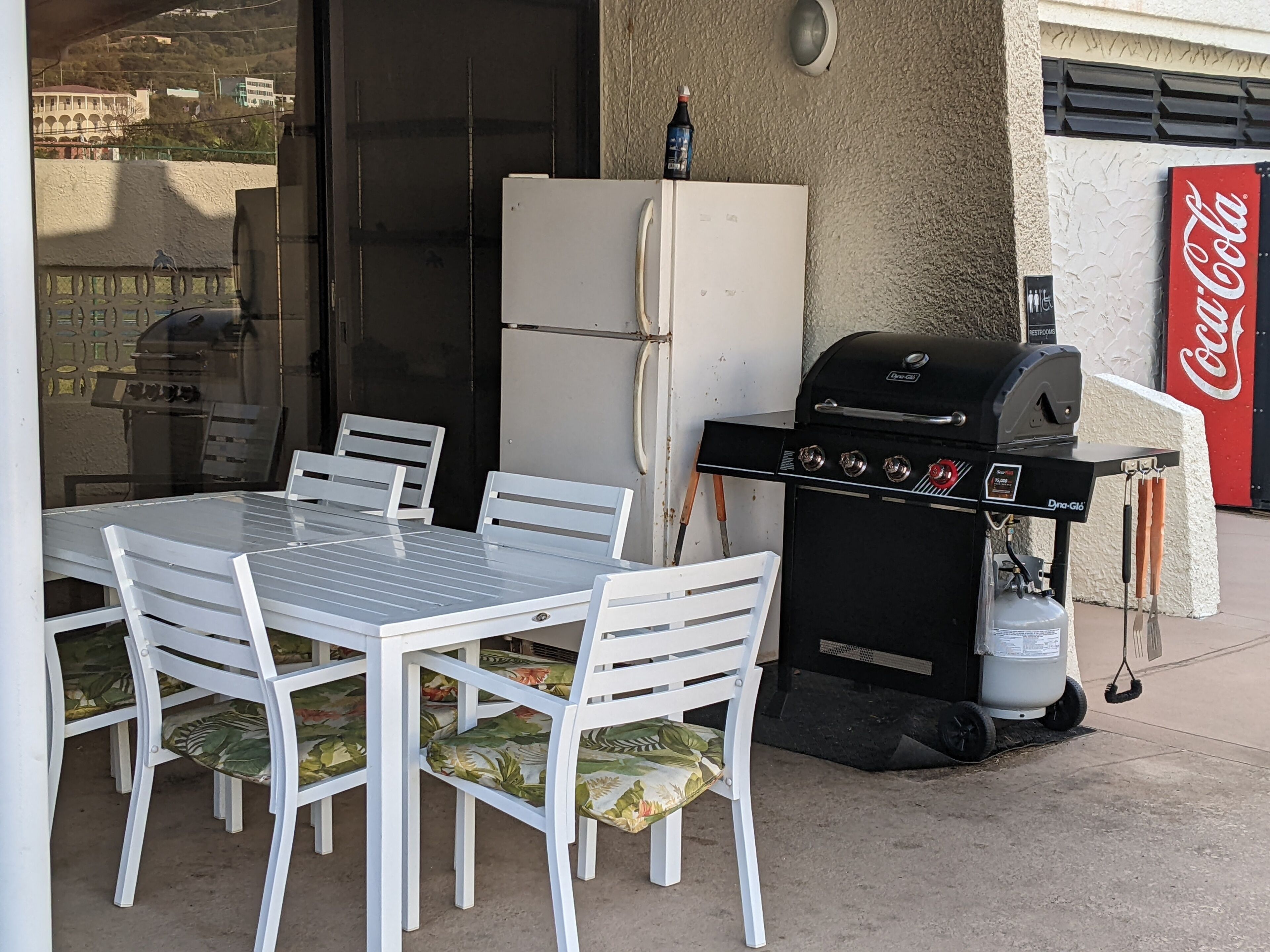 Een terras buiten met een tafel, stoelen, een barbecue, een koelkast en een Coca-Cola bord.