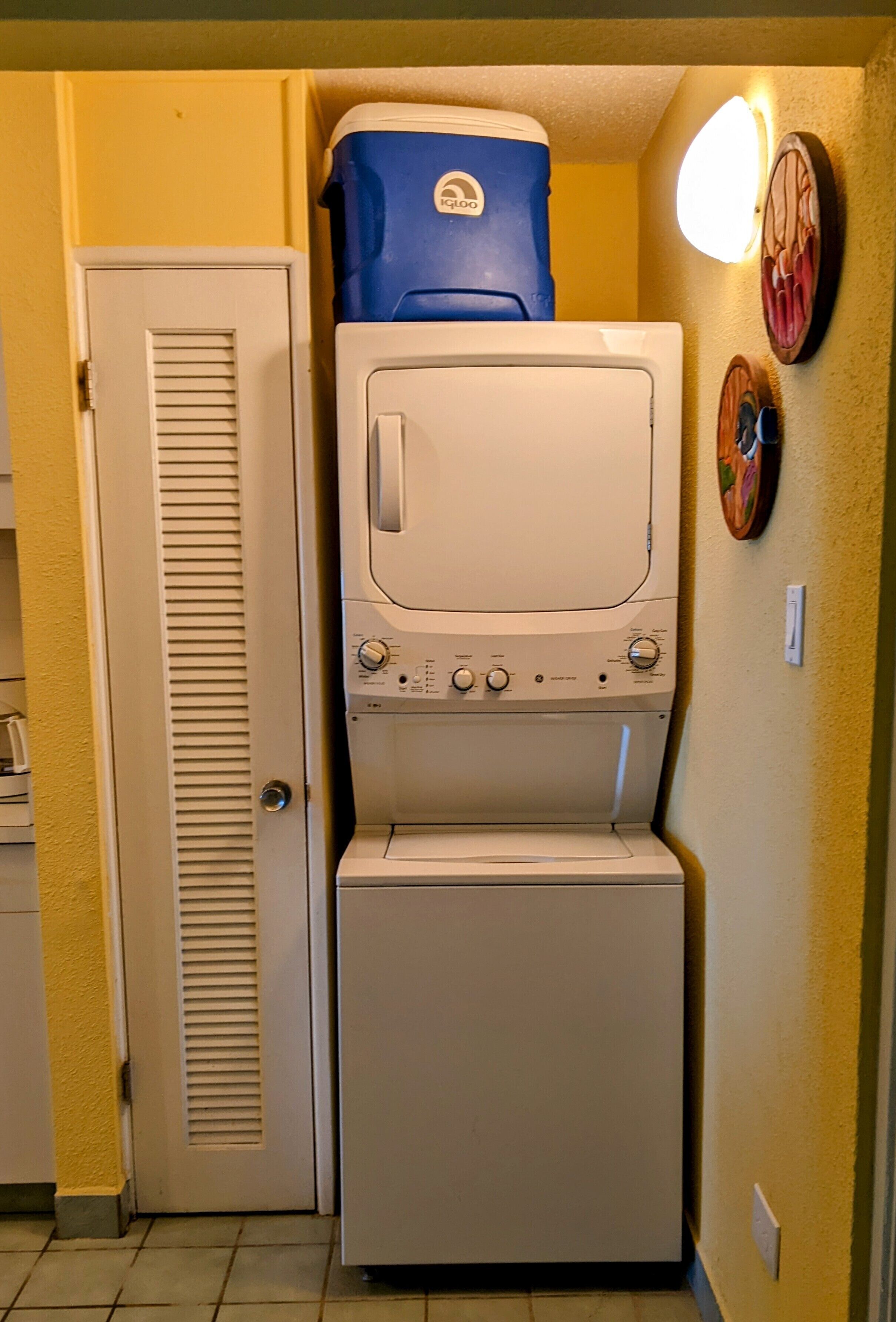 Une petite buanderie équipée d’un lave-linge et d’un sèche-linge à chargement frontal, d’un bac de rangement bleu posé sur le sèche-linge et d’une porte dotée d’une grille d’aération.