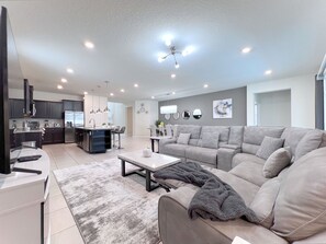 A modern living room with a grey sofa, a coffee table, and a kitchen area with dark cabinets and stainless steel appliances.