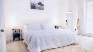 Une chambre à coucher avec un grand lit, deux tables de chevet, une commode, un miroir fixé au mur et une fenêtre avec des stores.
