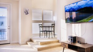 Un salon moderne avec un escalier, un téléviseur à écran plat fixé au mur, un bureau en bois avec un haut-parleur, et un coin repas avec une table et des chaises.