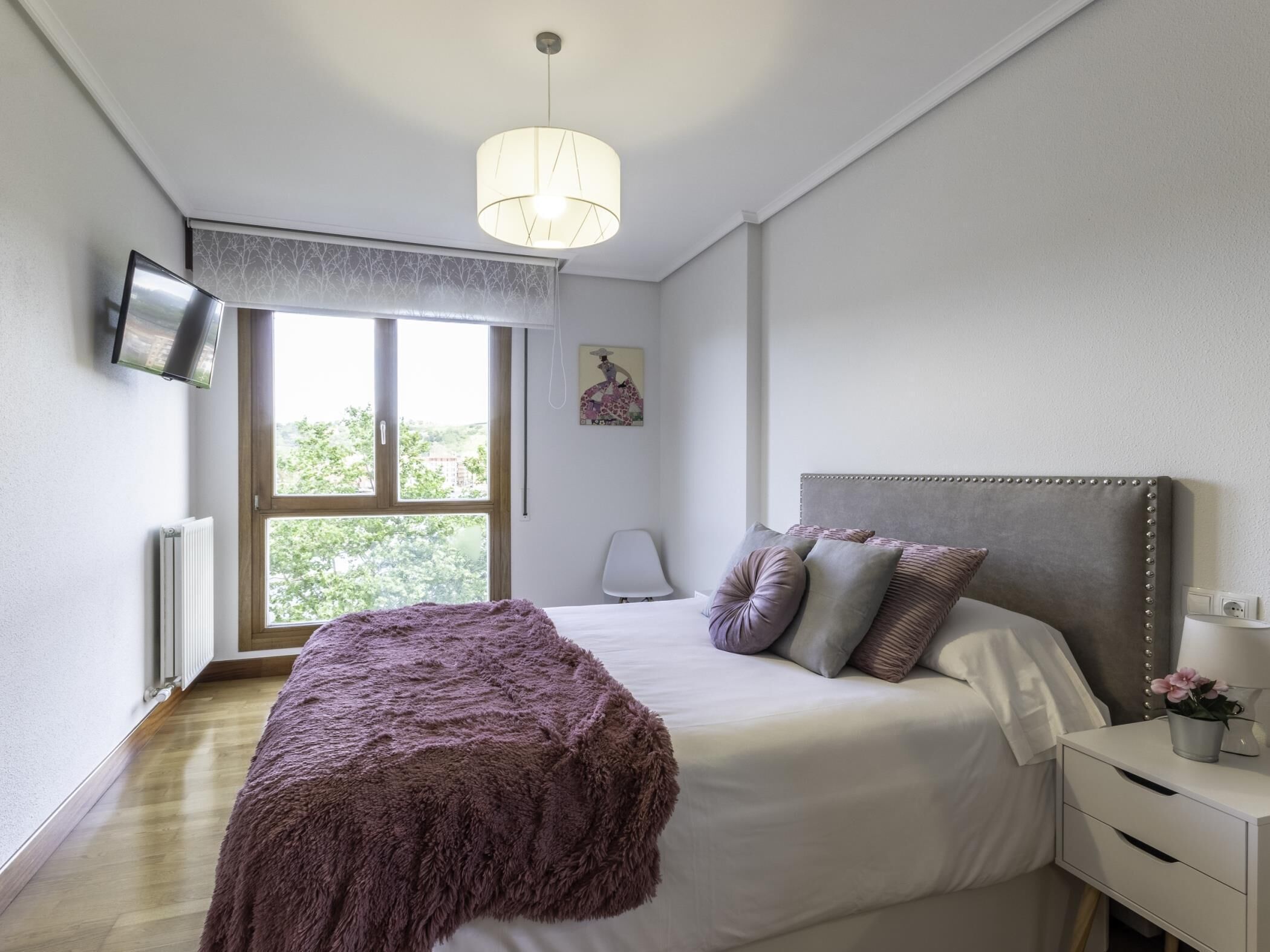 A bedroom with a bed, a bedside table with a lamp, a window with a view of trees, and a ceiling light.