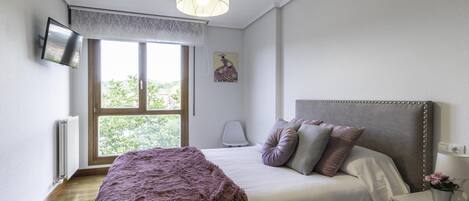 A bedroom with a bed, a bedside table with a lamp, a window with a view of trees, and a ceiling light.
