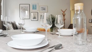 Mesa de jantar posta com pratos brancos, tigelas, copos de vinho e talheres. Quadros emoldurados estão na parede atrás da mesa.