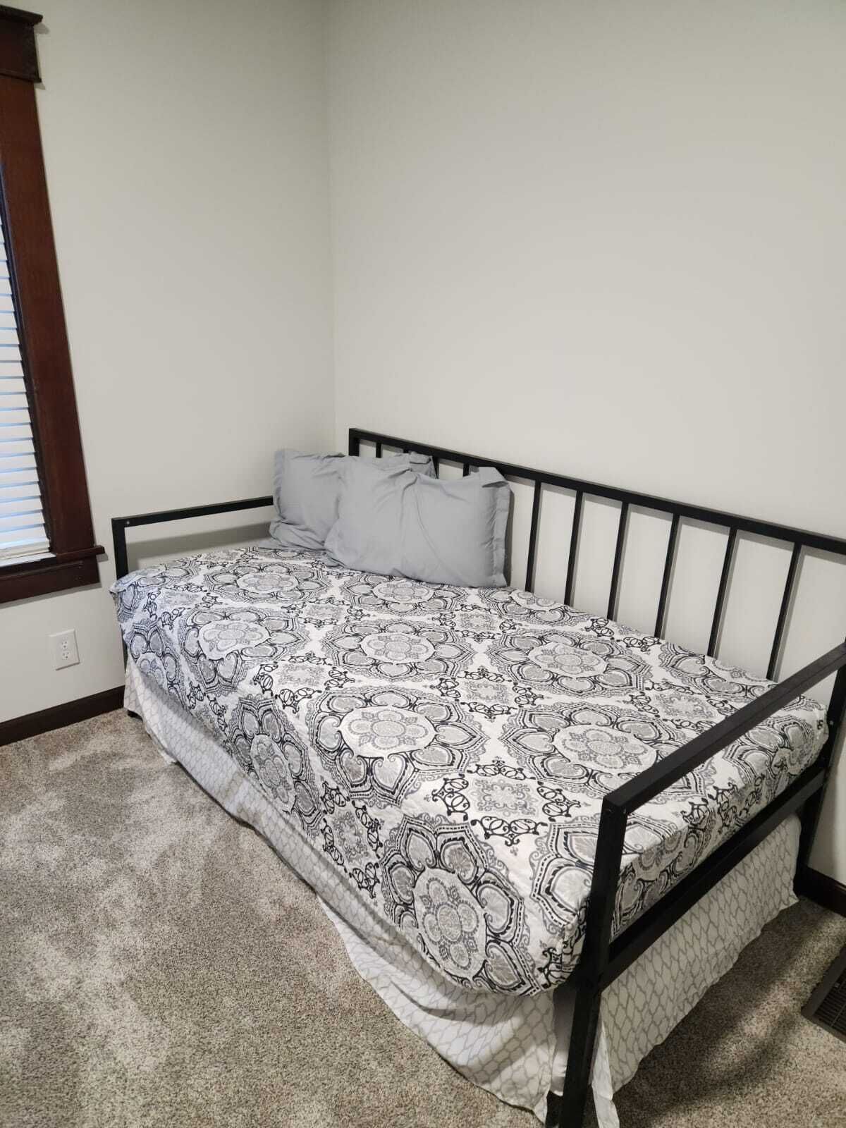 A single bed with a patterned bedspread and pillows, a wooden headboard, and a carpeted floor.