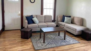 A living room with a beige sectional sofa, a wooden coffee table, a patterned rug, a clock on the wall, and a window with blinds. - Family Home in Carthage (Carthage)