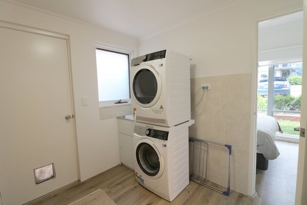 A compact laundry room with two stacked washing machines, a window, and a view of a car and building outside. - Beach Haven - 3 bedroom Fishpen (Merimbula)
