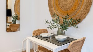 Un coin repas avec une table blanche, des chaises en osier, une décoration murale tissée et un miroir qui reflète une partie de la pièce.