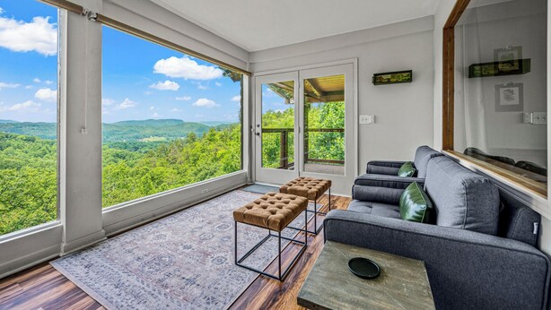 A spacious room with large windows offering a view of green hills and a clear sky. There is a gray sofa and two tufted ottomans. - 🌟Private🌟Views🌟x2KingSuites🌟HotTub🌟FirePit🌟NationalPark🌟SunSet🌟 (Sevierville)