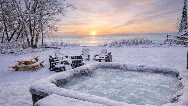 A snow-covered outdoor hot tub with chairs and a picnic table, surrounded by trees and a view of the sunset over the water. - Lakefront Cabin Hot Tub Playground Fire Pit (Algoma)