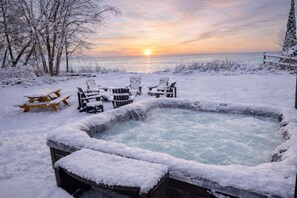 A snow-covered outdoor hot tub with chairs and a picnic table, surrounded by trees and a view of the sunset over the water. - Lakefront Cabin Hot Tub Playground Fire Pit (Algoma)