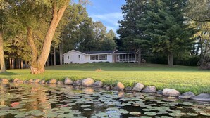 A pond with lily pads and a house with a lawn in the background. - Private, newly renovated lakefront cottage on 10+ acres of land. (Brooklyn)