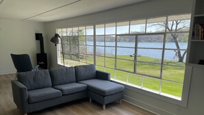 A modern living room with a grey sectional sofa, a black floor lamp, and a large window overlooking a lake. - Private, newly renovated lakefront cottage on 10+ acres of land. (Brooklyn)