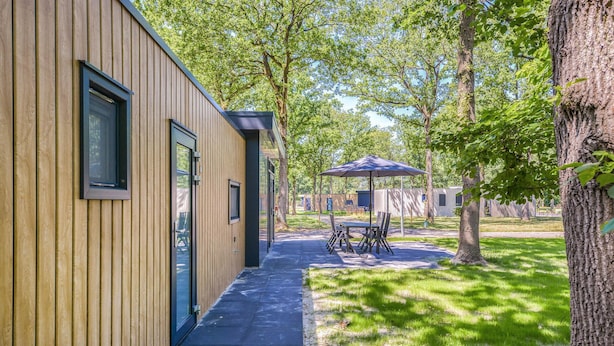 Een modern gebouw met een houten gevel, een overdekt terras met tuinmeubilair en een gazon met bomen.