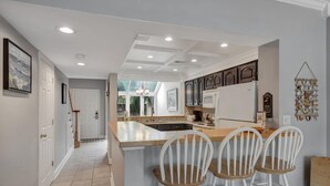 A kitchen with a central island, white bar stools, dark wooden cabinets, and a view to the outdoors. - Beachwalk Villa Retreat w/ Golf Cart & Pool Access (Miramar Beach)