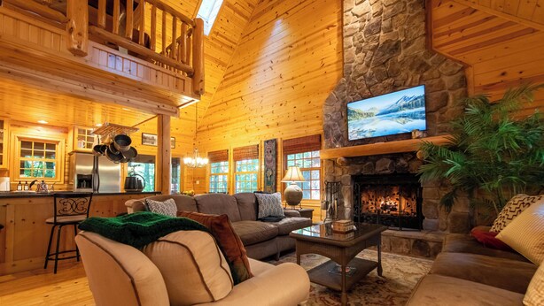 A log cabin interior with a stone fireplace, wooden ceiling, and a cozy living area with sofas and a television.
