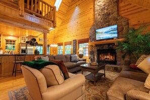 A log cabin interior with a stone fireplace, wooden ceiling, and a cozy living area with sofas and a television. - Log cabin with private hot tub and shared pool access! (Saugatuck)