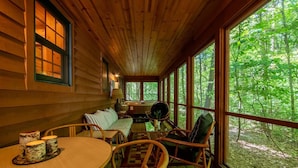 A cozy cabin interior with a wooden ceiling, large windows offering a forest view, a round dining table with chairs, and a sofa with a view.