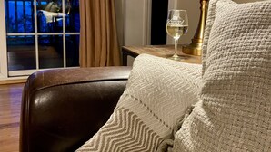 Een knus ingerichte kamer met een leren fauteuil, een geweven sierkussen, een messing vloerlamp en een glas wijn op een bijzettafeltje.