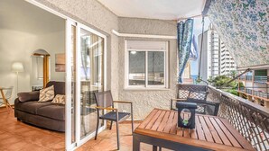 A balcony with a wooden table and chairs, a sofa, and a sliding glass door leading to an indoor living area with a lamp and a chair. - Lovely apartment in Campello (Campello)