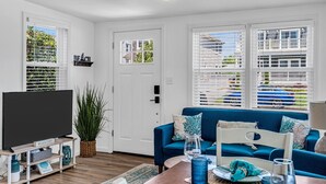 A living room with a blue sofa, a flat-screen TV, and a large window with blinds. - THE Unique Belmar Stay | Prime Location. Privacy. 9 Badges. 2 Grills. Fire Pit. (Belmar)