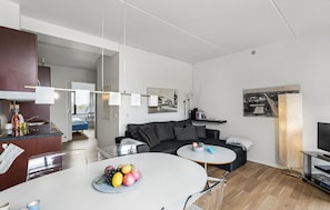 A modern living room with a grey sofa, a round coffee table with fruit, a TV stand, and a dining area with a white table. - Beautiful apartment in Fanø with WiFi (Fanø)