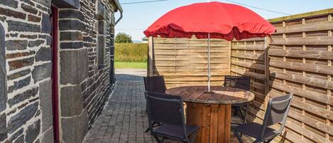 A stone building with a red door, a wooden table with four chairs, and a red umbrella.