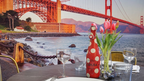 Ein Esstisch, gedeckt mit einer Flasche Lolea Wein, einem Glas und einer Vase mit Tulpen, mit Blick auf die Küste und einer Brücke im Hintergrund.