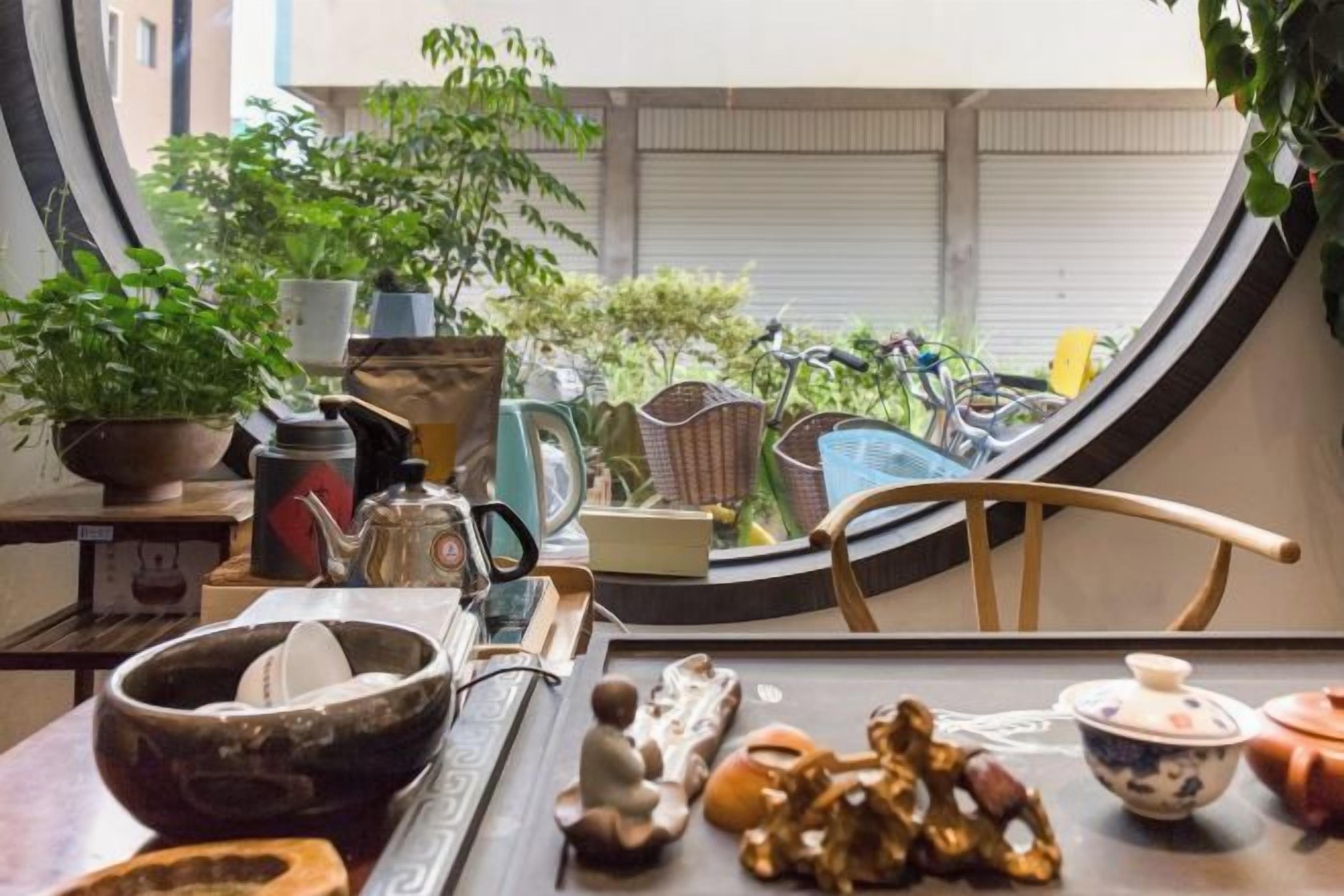 Uitzicht door een raam, met een tafel, stoelen en diverse spullen, waaronder een kom, een theepot en een bloempot.