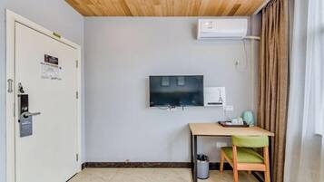 A hotel room with a wooden ceiling, a flat-screen TV mounted on the wall, a desk with a chair, and an air conditioning unit.