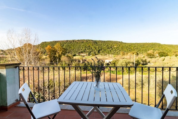 A balcony with a table and two chairs, overlooking a hilly landscape with trees and a clear sky.
