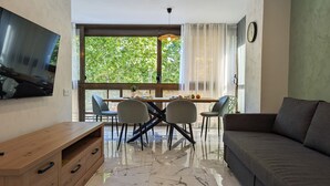 A modern living room with a sofa, a TV mounted on the wall, a dining table with chairs, and a large window with a view of trees. - LAS OLAS old town apartments (Benidorm)