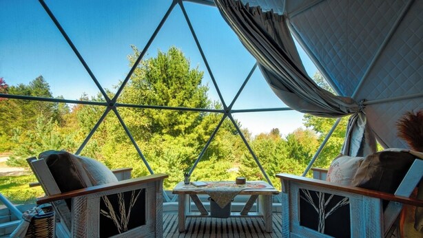 A geodesic dome structure with a wooden floor, two chairs, a table, and a view of trees and greenery outside. - Luxury on the Edge of Traverse City - 6 (Williamsburg)