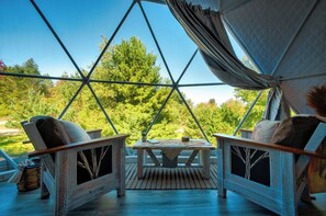 A geodesic dome structure with a wooden floor, two chairs, a table, and a view of trees and greenery outside. - Luxury on the Edge of Traverse City - 6 (Williamsburg)